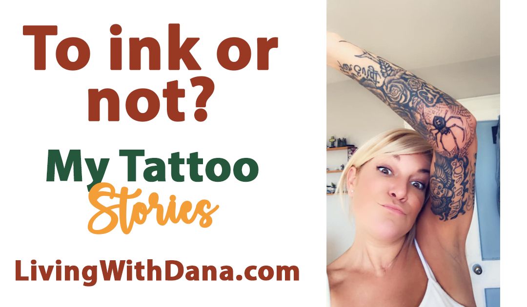 A woman playfully lifts her arm to show detailed tattoos on her inner arm, including a spider and floral designs. She’s smiling and expressive in a white tank top indoors.