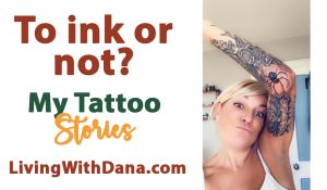 A woman playfully lifts her arm to show detailed tattoos on her inner arm, including a spider and floral designs. She’s smiling and expressive in a white tank top indoors.